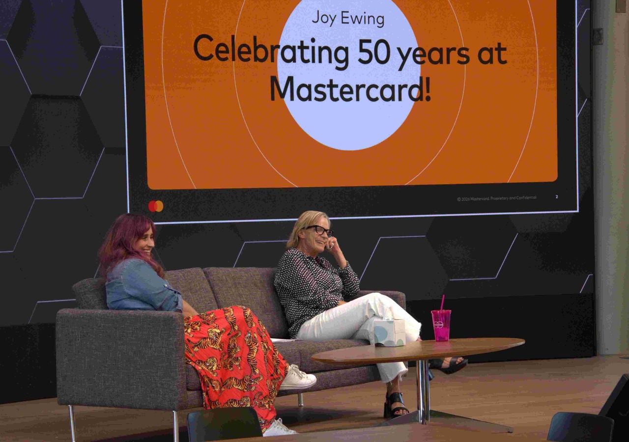 Shannon Burke-Kranzberg and Joy Ewing sit on a couch in front of a sign celebrating Joy's 50 years at Mastercard. 