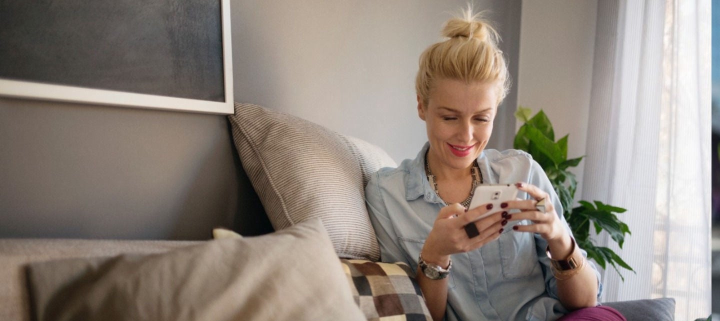 A woman is seated on a couch in her home smiling at her phone.