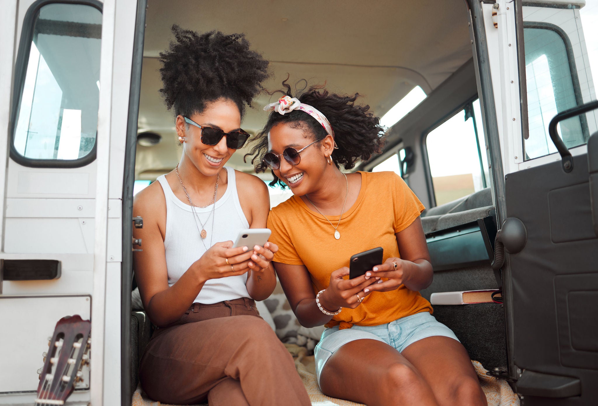 Two smiling young women sitting in the back of a van on a road trip, laughing and looking at their smartphones.
