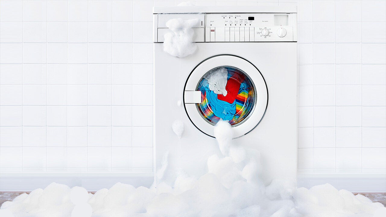 Túlhabzó mosógép színes ruhákkal, kifolyó mosóhabbal fehér csempézett fürdőszobában
