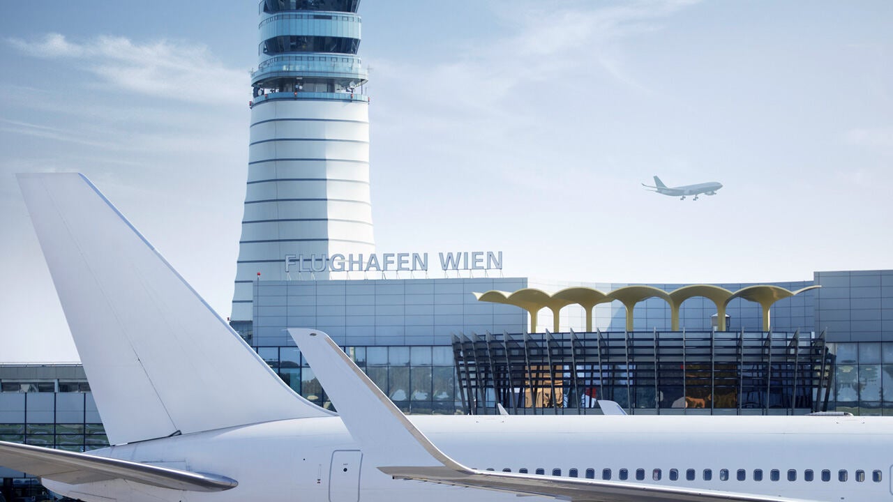 Repülőtér terminálépülete irányítótoronnyal, előtérben utasszállító repülőgép szárnya, háttérben felszálló repülőgép