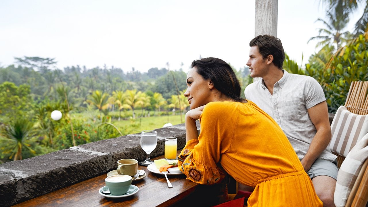 A couple dressed in casual wear sit on a patio eating breakfast while looking out at a beautiful tropical vista. 
