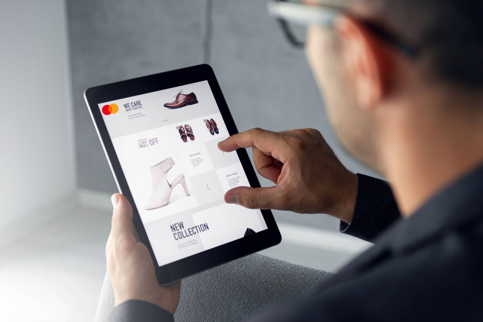 An over-the-shoulder view of a man in glasses tapping a tablet displaying an online shoe shopping page.