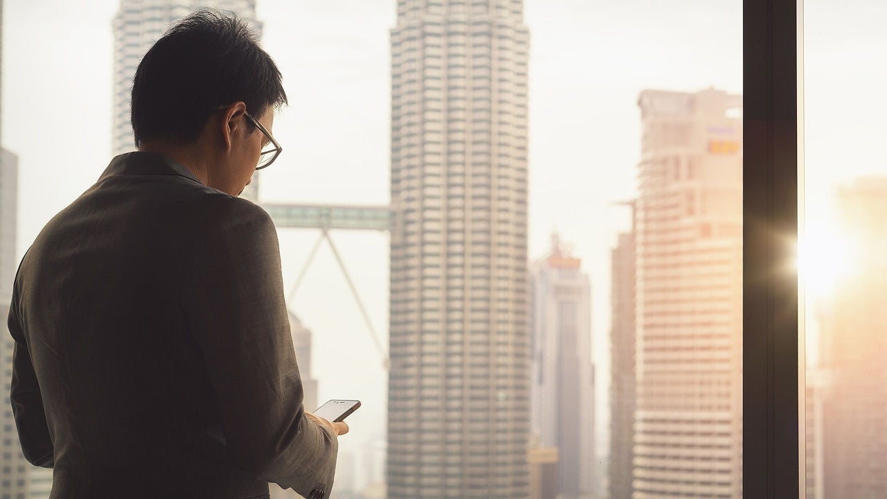 Man in glasses looking at his phone beside a window overlooking a city skyline.