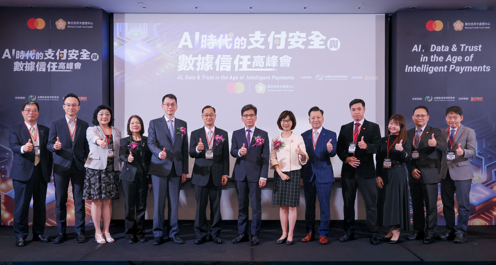 A group of thirteen people in business attire stand on a stage giving thumbs up in front of a large screen at a conference.