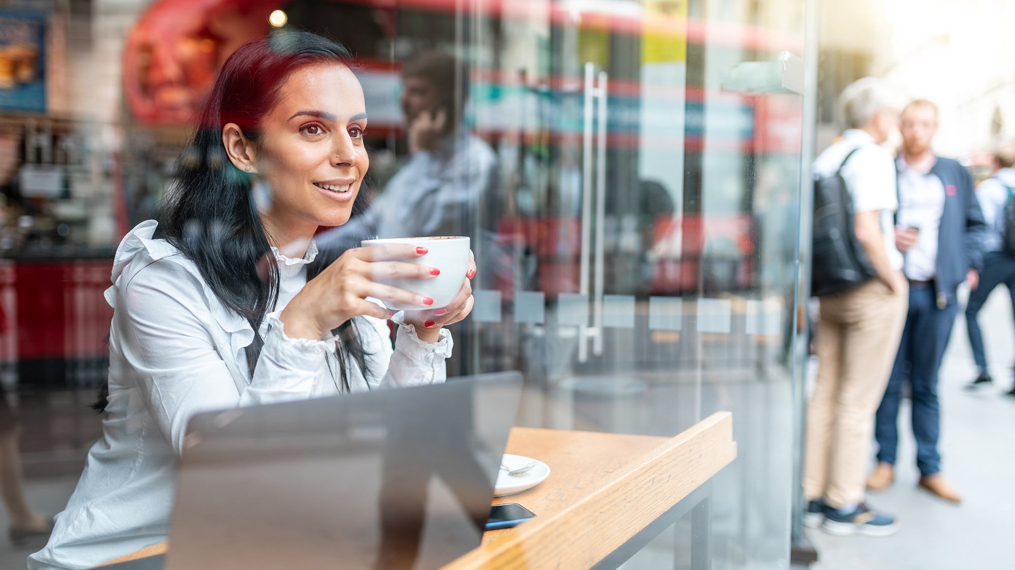 Professional woman smiling, drinking coffee, and working on a laptop by a cafe window, with reflections of a city street and a red double-decker bus.