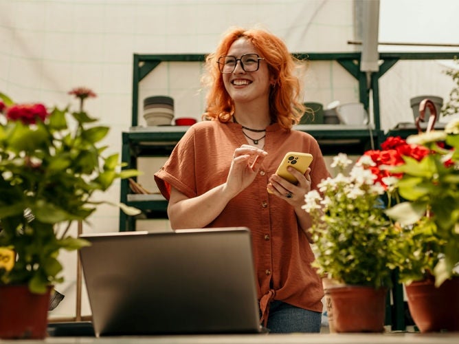 A woman holds a phone and looks over a laptop in a garden shop. 