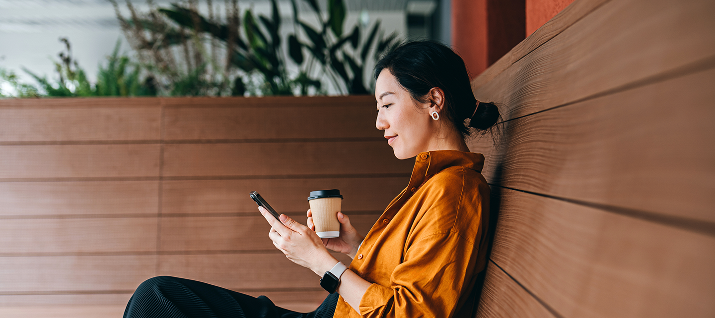 Young asian woman sitting in a sidewalk cafe with a cup of coffee looking at her phone