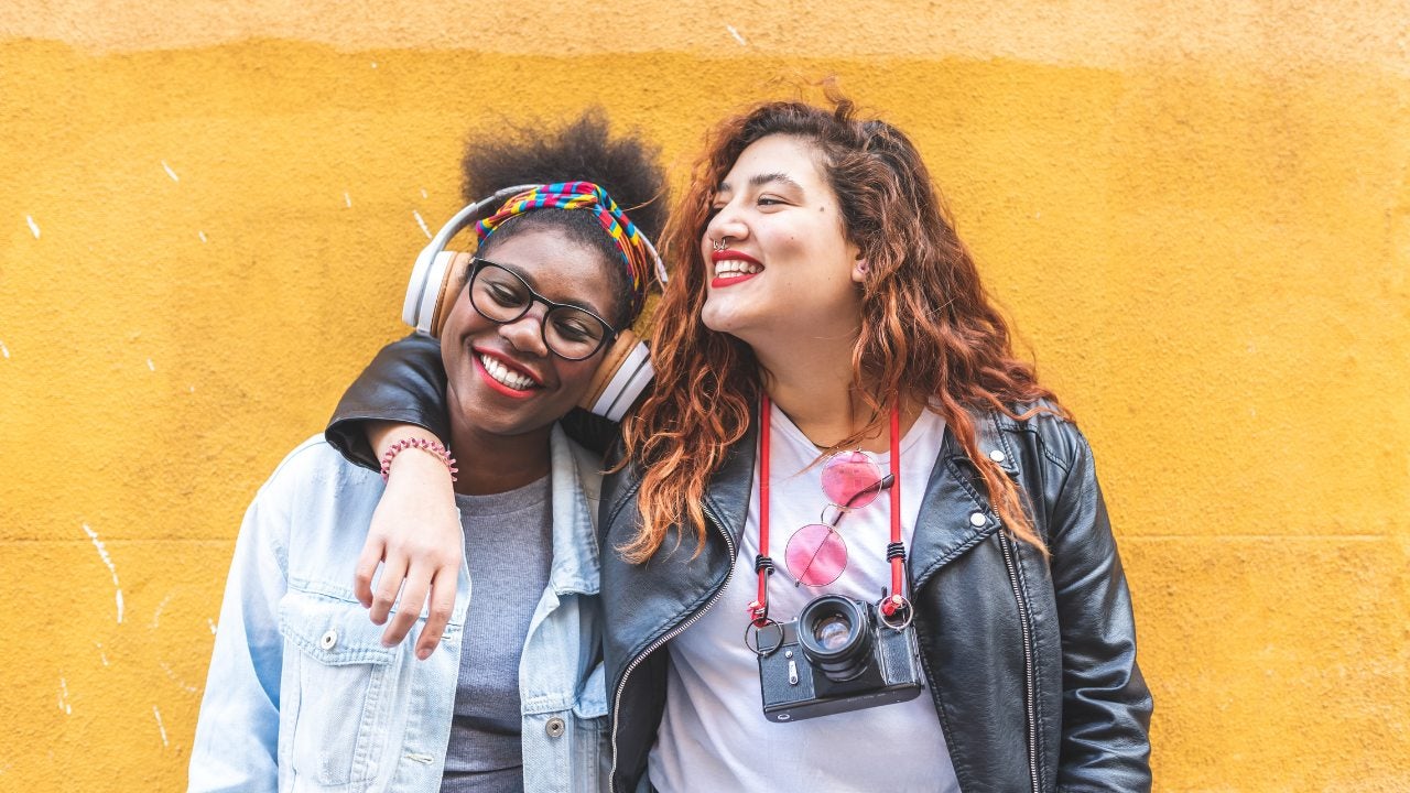 Two young woman, one listening to headphones, the other with a camera on a strap around her neck, stand together with arms around each other and smile.