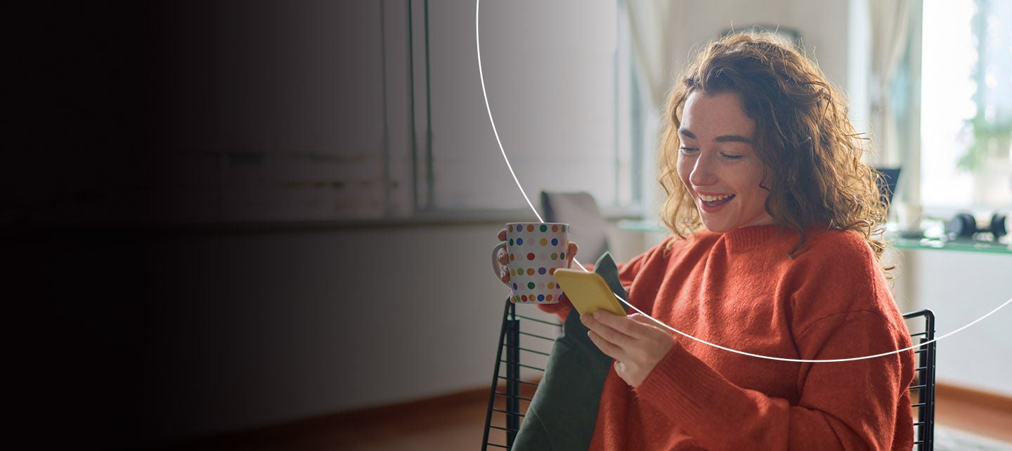 žena sa šoljom kafe koristi telefon u kuhinji.