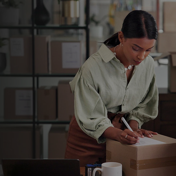 A woman in a light green blouse seals a cardboard box with packing tape while working at a table in a storage room with stacked boxes in the background.