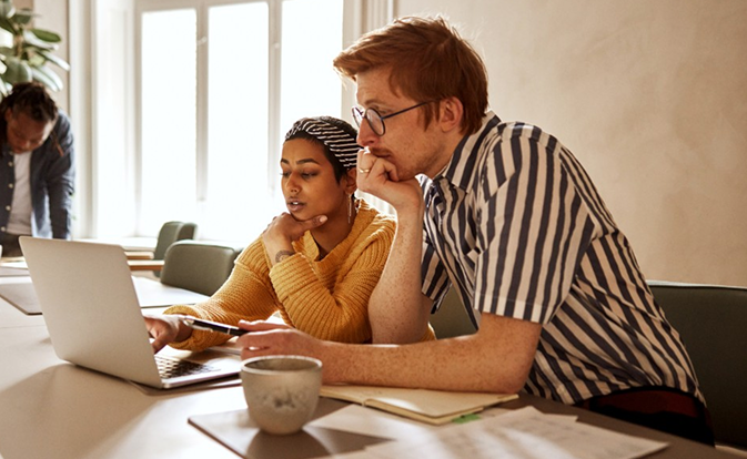 Diverse business professionals, a woman and a man, collaborating intently on a laptop in a brightly lit modern office, showcasing teamwork and discussion.