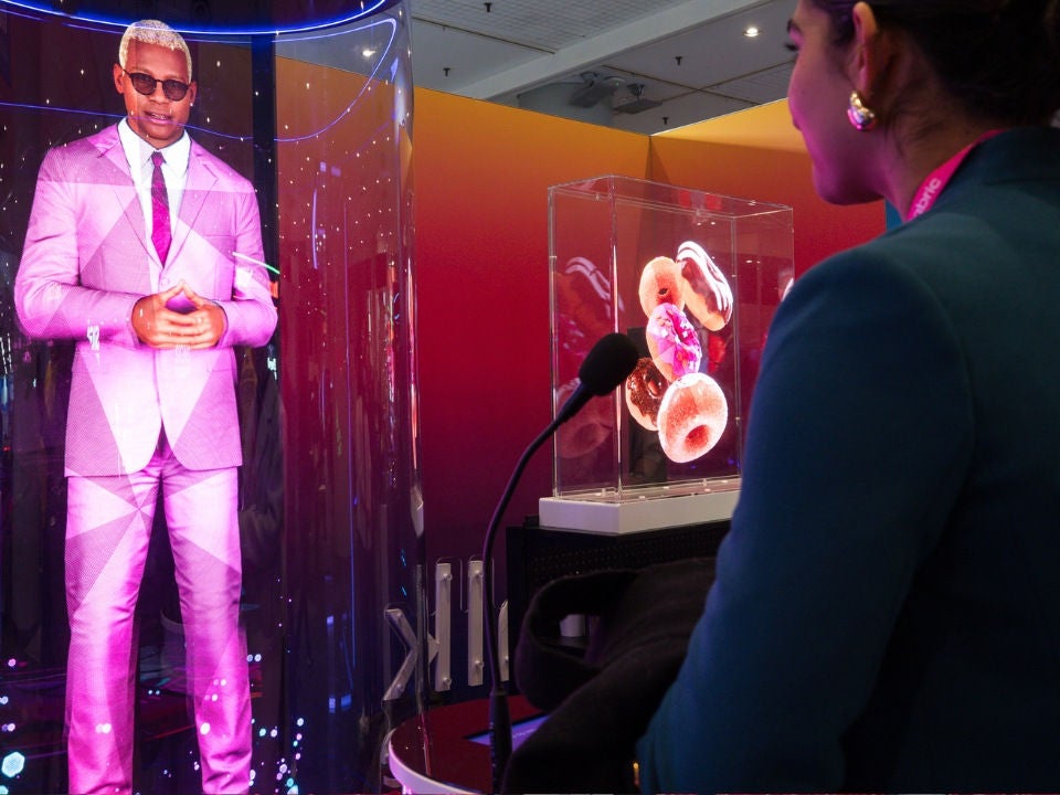 An NRF attendee stands in front of a microphone in front of a hologram of a stylishly-dressed man in sunglasses. 