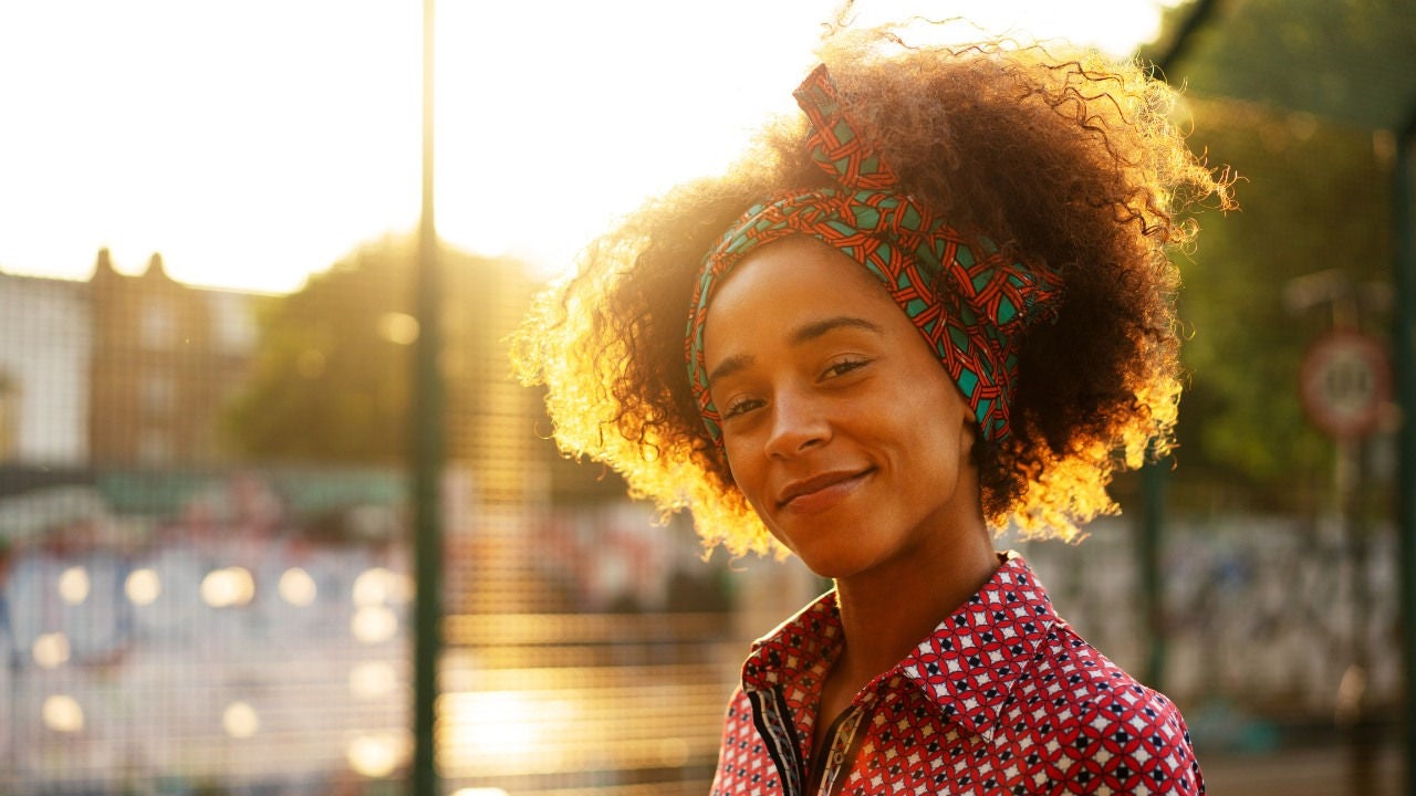 A woman wearing a patterned piece of fabric around her head smiles in the setting sun. 