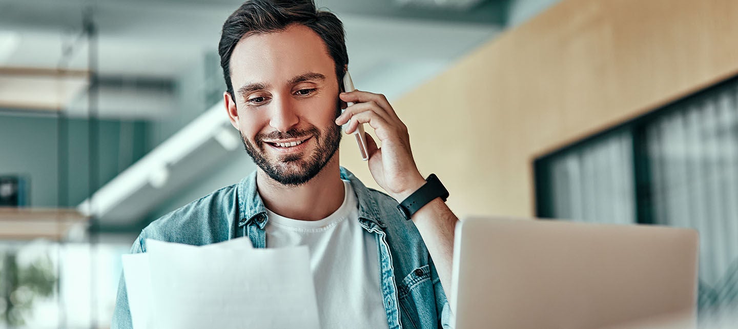 Young man on the phone holding financial papers while working on a laptop