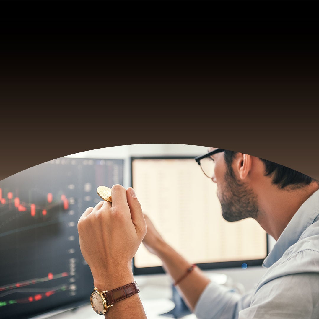 Man at a desk holding a coin, analyzing financial data on dual monitors showing a candlestick chart and numerical metrics.