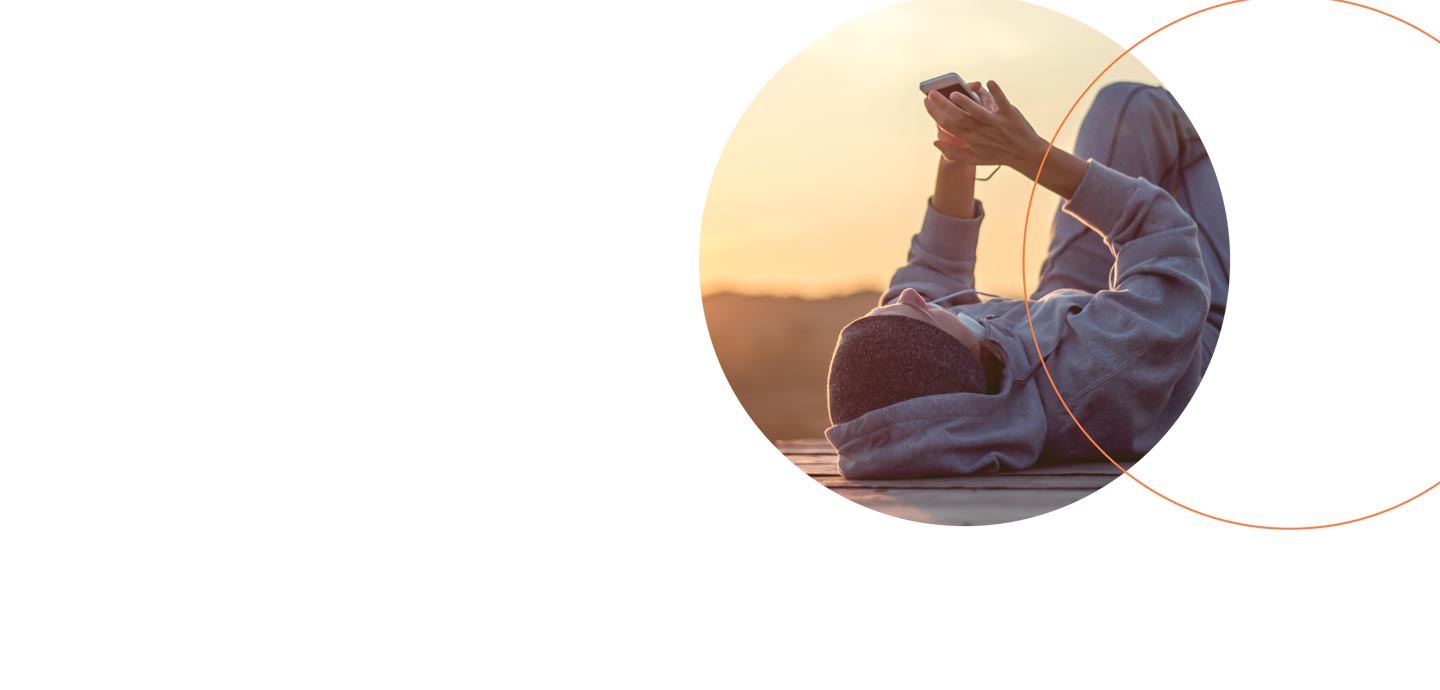 En person ligger på ryggen udendørs og holder en smartphone op foran sig. Personen har en hættetrøje og hovedtelefoner på, og baggrunden viser en solnedgang.