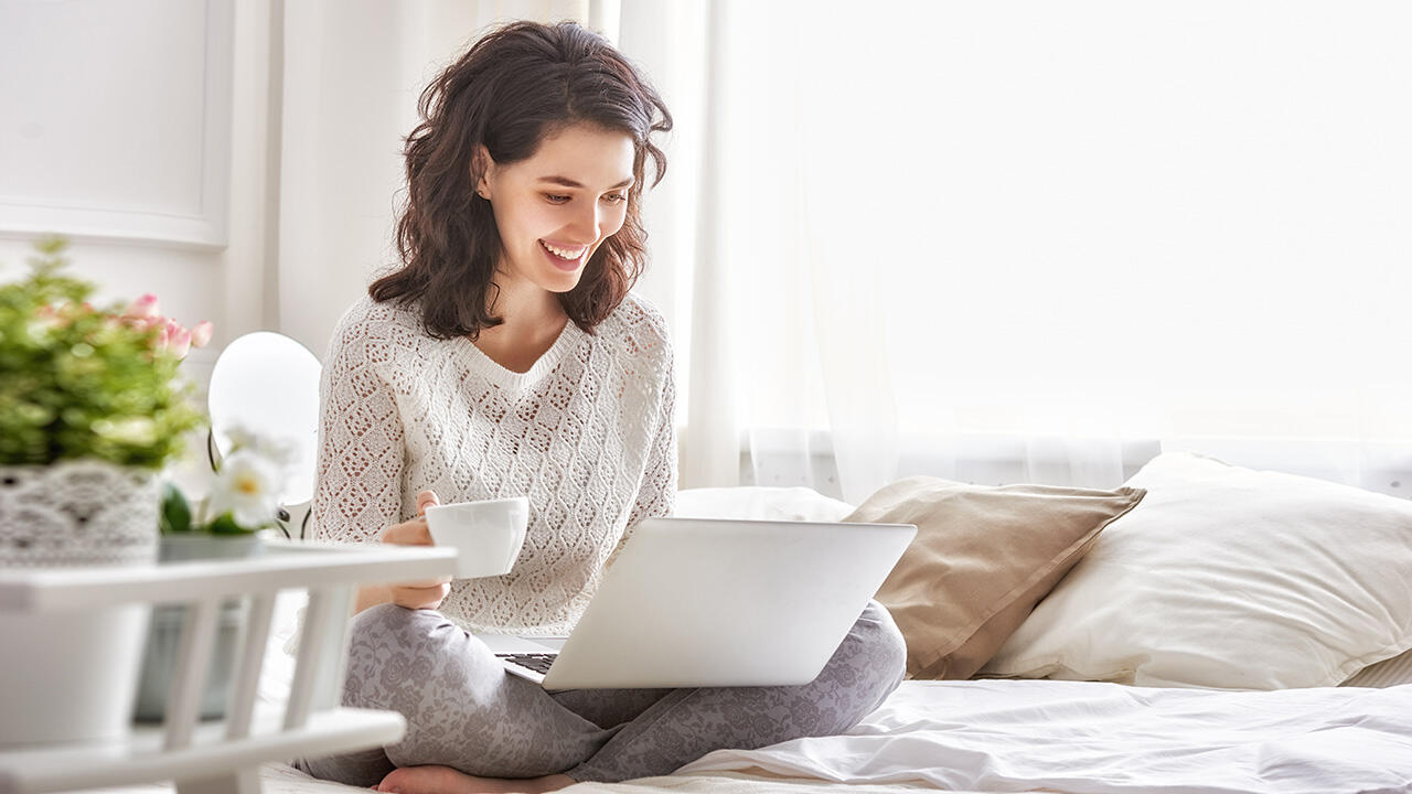 Mosolygó nő otthon, ágyon ülve laptopon dolgozik vagy böngészik, miközben egy csésze kávét tart a kezében.