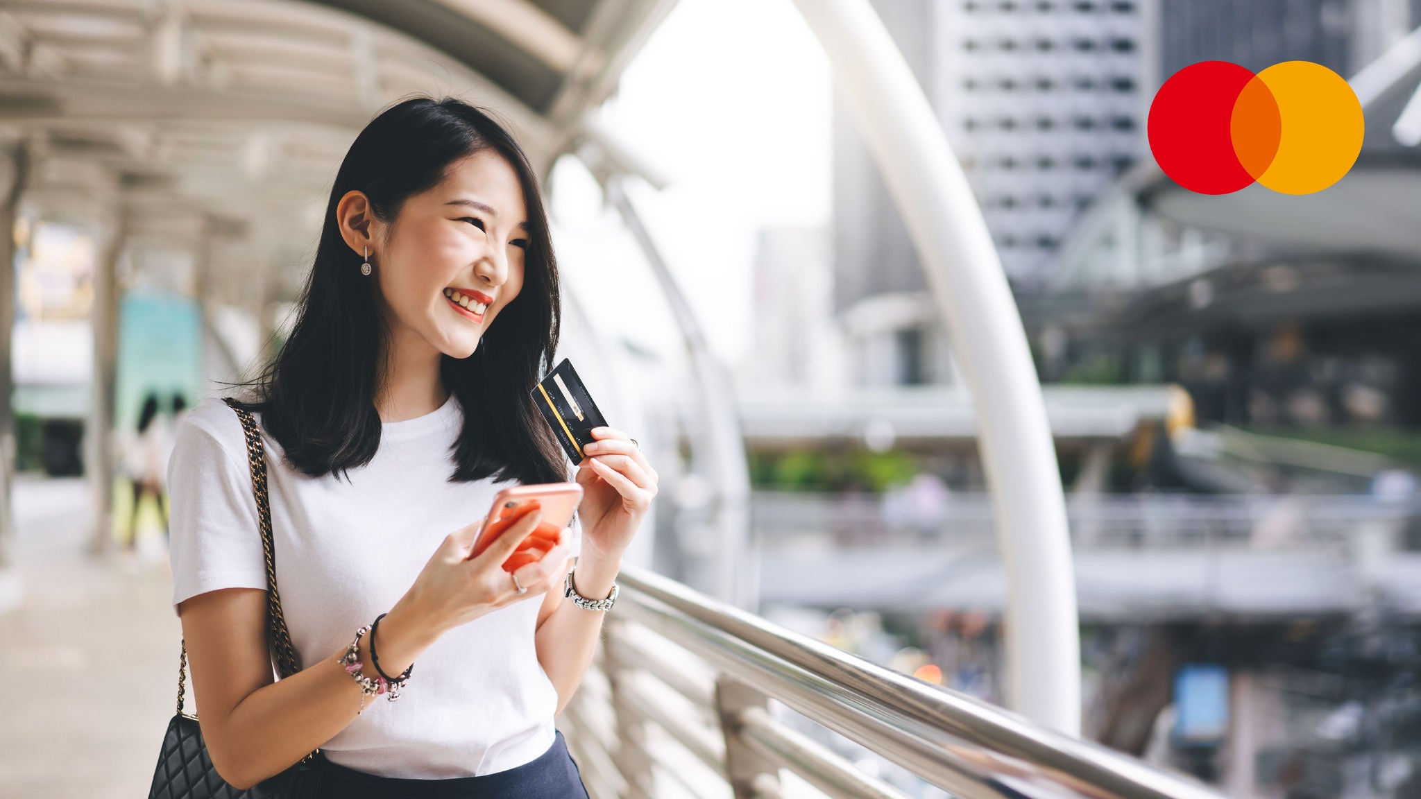 A woman stands on an outdoor city walkway holding a credit card and a mobile phone, smiling as she looks into the distance.