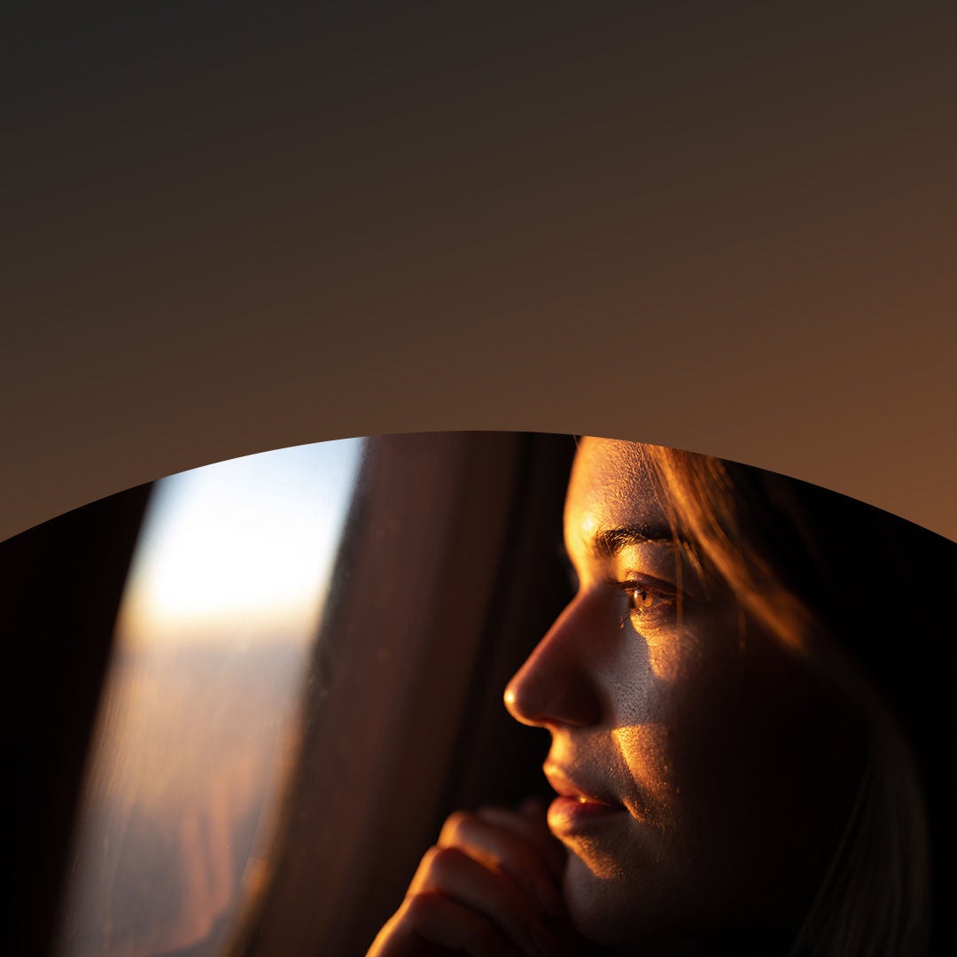 A woman gazes out the airplane window, observing the clouds and landscape below during her flight.