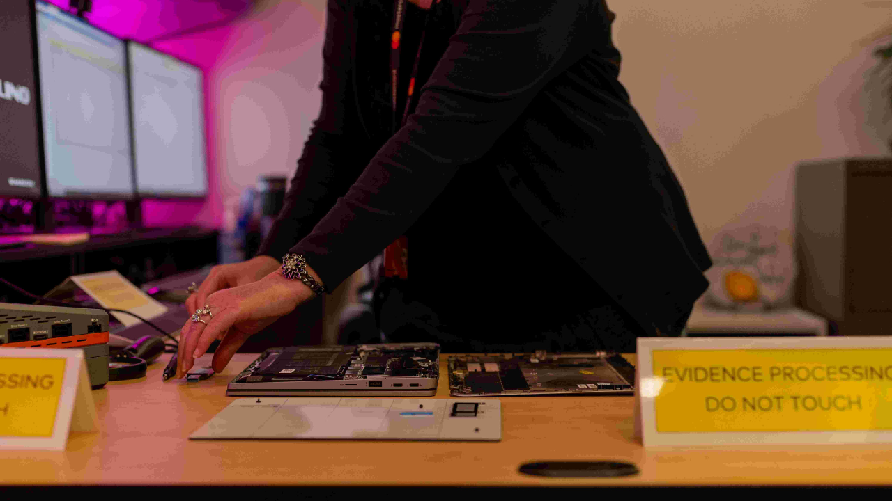 A woman handles the inside of a computer on a table that says "evidence processing, do not touch." 