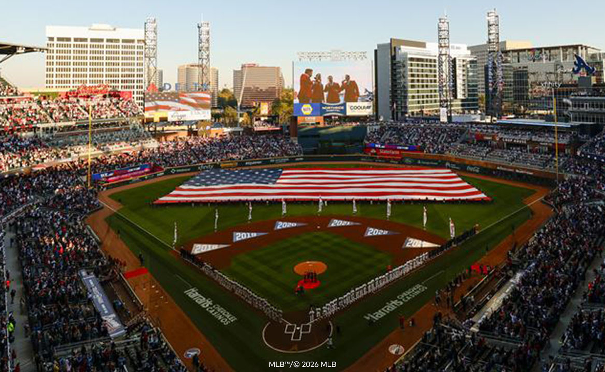 An aerial view of a packed baseball stadium with a large American flag on the field, evoking priceless moments at the game.