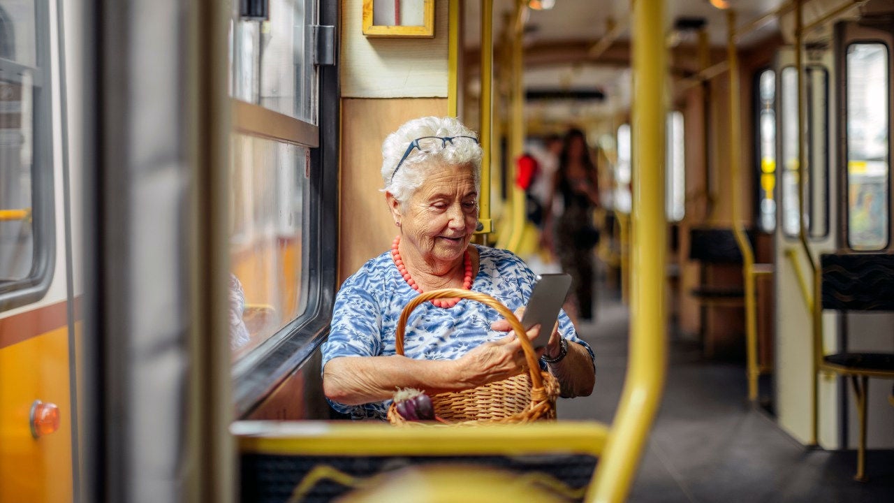 An older woman on a train holds a basket in her lap while look at a cell phone. 