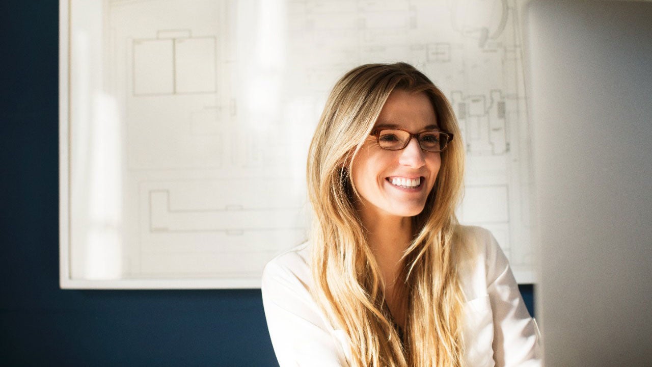 En kvinde med lyst hår og briller smiler glad foran en tegning på en væg.