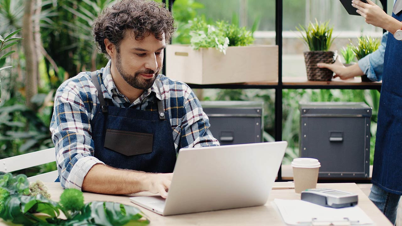 Man typing on laptop at desk in greenhouse.