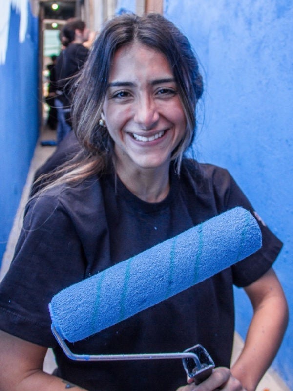 Beatriz Mello holds a paint roller in front of a house face she is painting. 