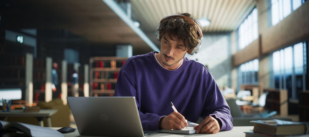 Young man in library wearing headphones, taking notes while studying on a laptop 