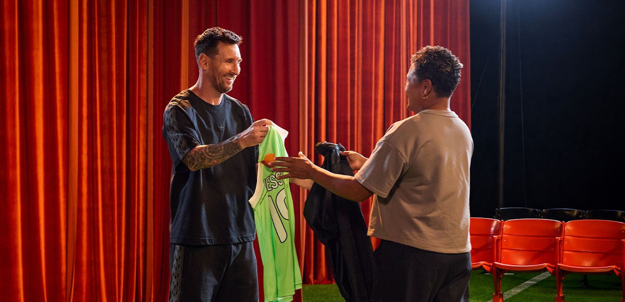 Lionel Messi smiling as he exchanges jerseys with a female fan. Messi holds a bright green, signed "Messi 10" MasterCard jersey, while the fan hands over a dark shirt on a stage with a red curtain backdrop.