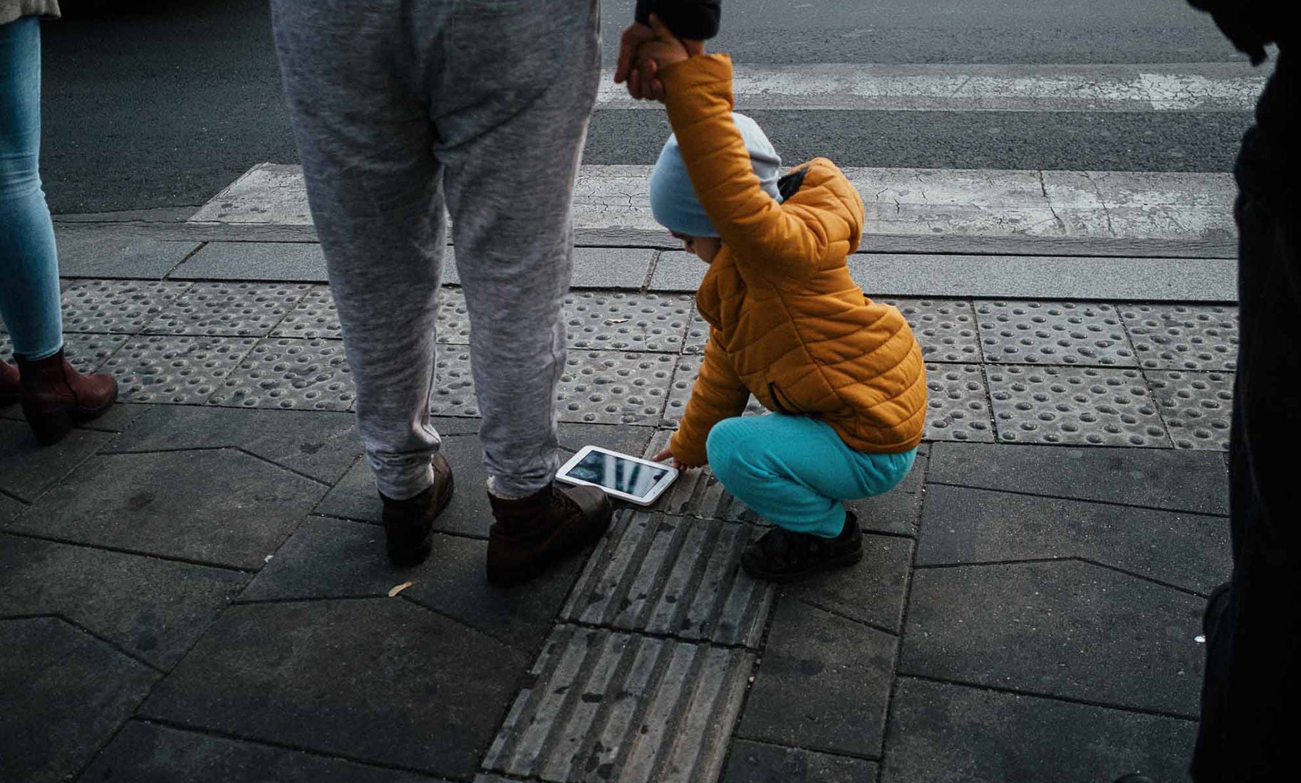 Egy kisgyermek leguggol egy zebránál elejtett táblagéphez, miközben egy felnőtt fogja a kezét.