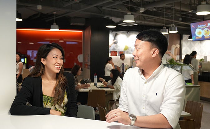 Two coworkers talking at a table in a casual office setting.