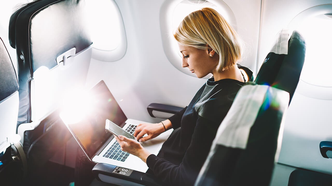 Fiatal nő repülőgépen ülve laptopon dolgozik és okostelefont használ