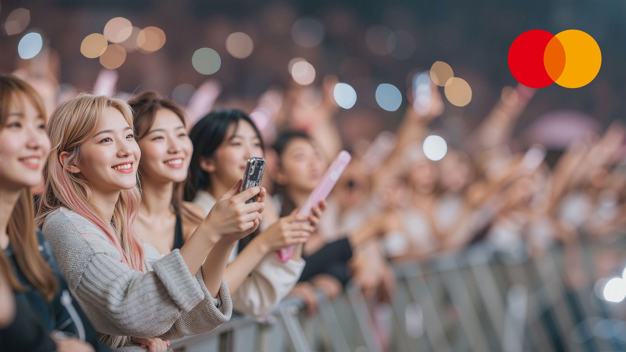 Fans filming a concert on their phones; Mastercard logo at upper right.