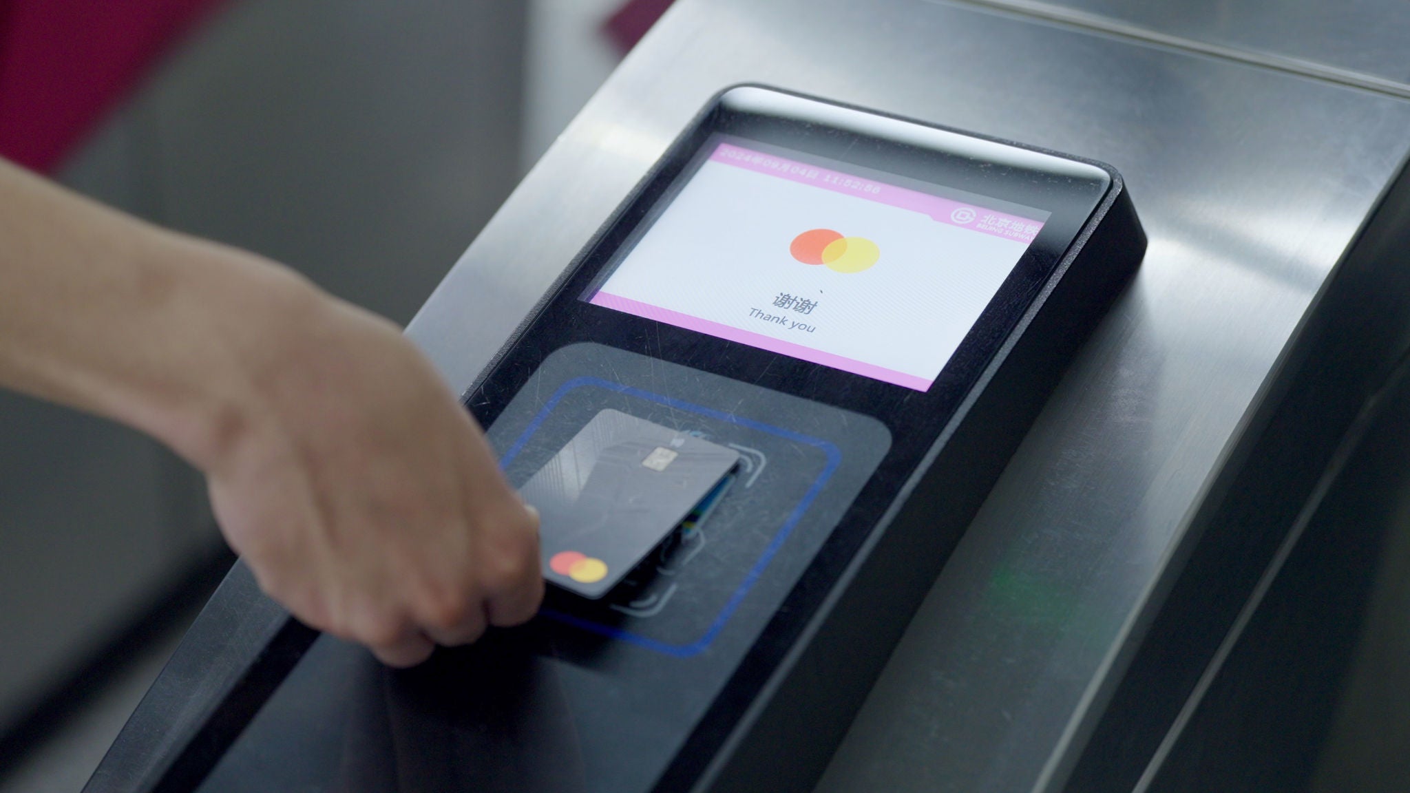 A hand taps a dark payment card on a subway gate reader, with the screen displaying a thank you message.
