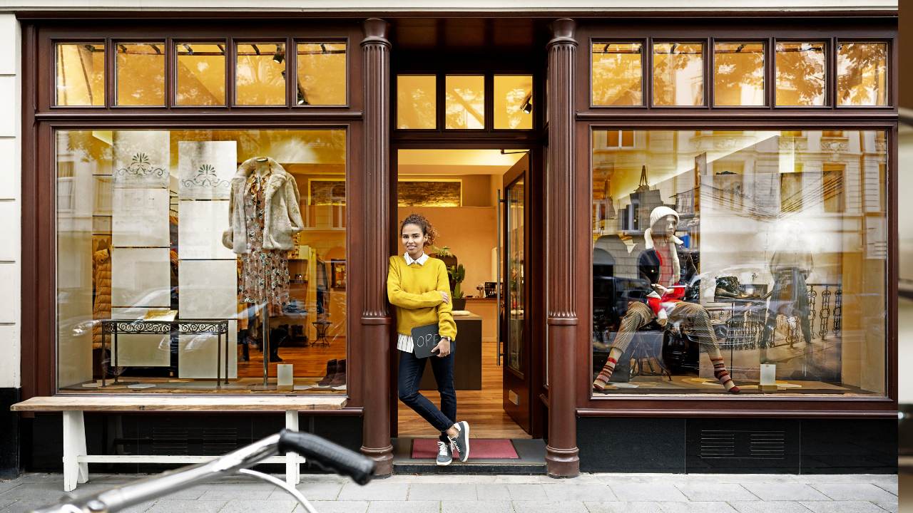 A store owners stands in front of her store holding an open sign.