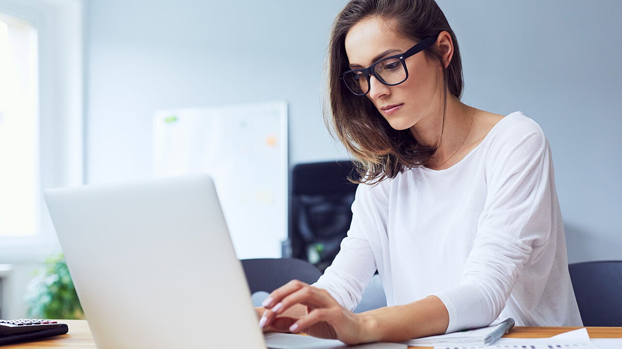 Irodában dolgozó nő laptopon gépel, szemüveget visel, az asztalon dokumentumok és toll látható