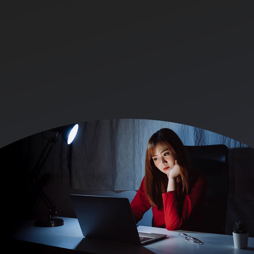 Woman in red shirt working on a laptop at a dimly lit desk with a lamp