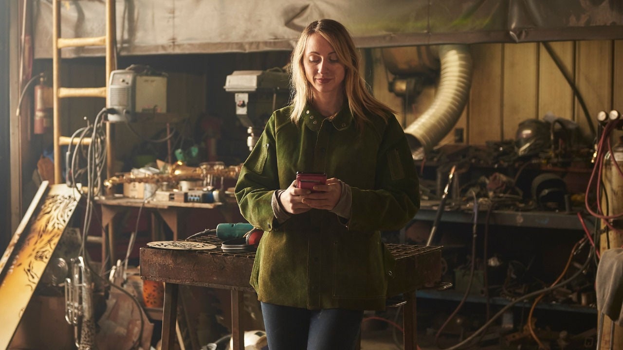 A woman in a heavy work coat looks at her phone in a workshop. 