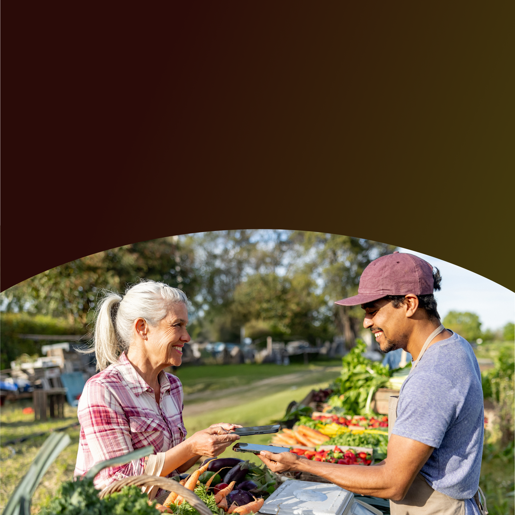 A smiling customer uses her smartphone to make a contactless mobile payment to a vendor holding his phone at an outdoor farmers market stand filled with fresh produce.