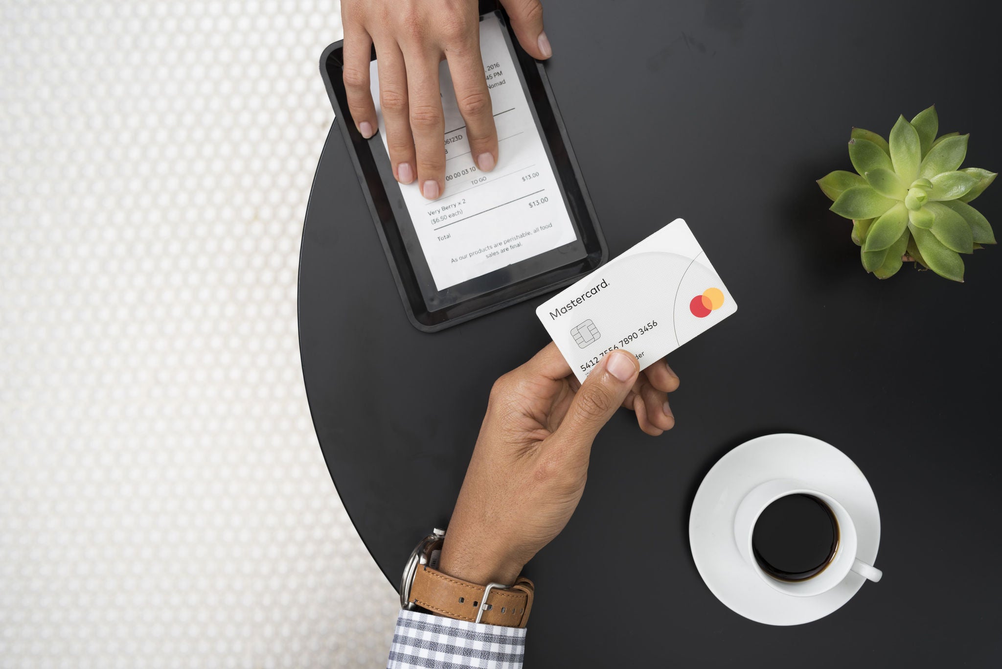 An overhead view of a hand holding a white payment card beside a black tray with a printed receipt, a cup of coffee, and a small succulent.