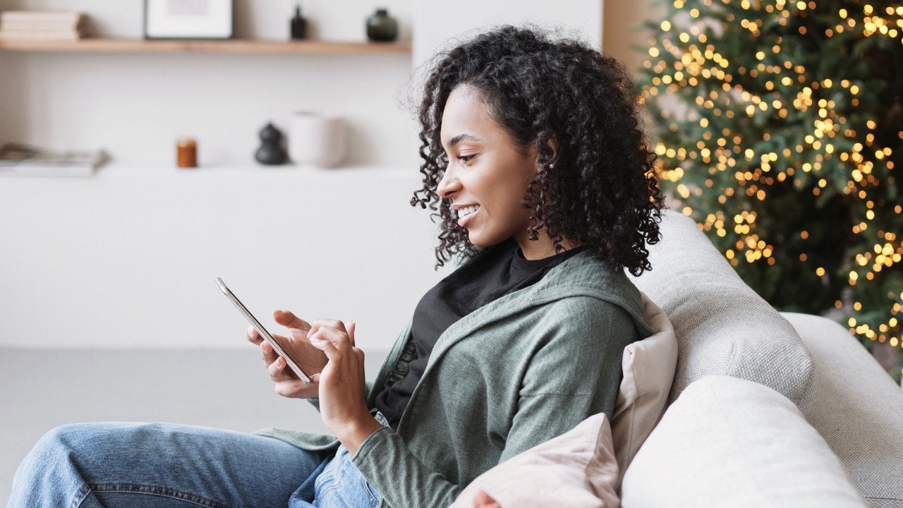 A woman sits on a couch looking at her phone in front of a decorated Christmas tree. 