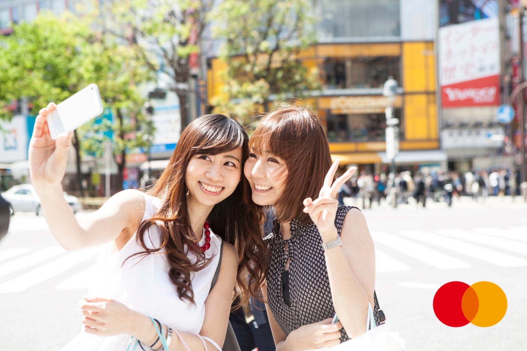 Two young women take a selfie together on a sunny city street, one holding up a phone.