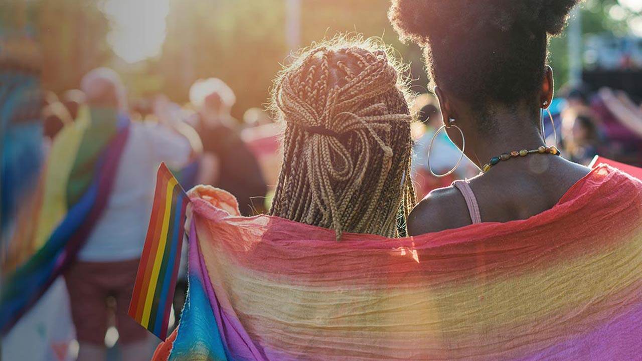 Két ember hátulról látható egy szabadtéri felvonuláson, vállukon szivárványszínű zászlóval, napfényben, sokszínű tömegben.