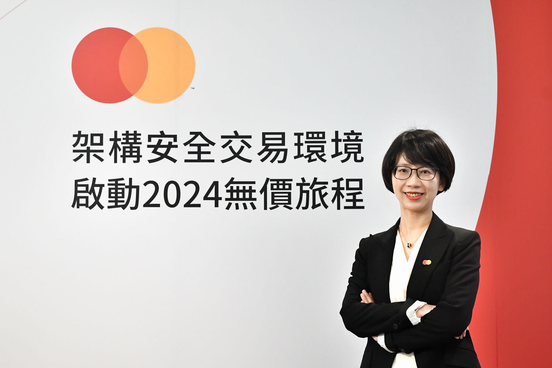 A woman in a black blazer stands with her arms crossed, smiling in front of a white and red backdrop with Chinese text and a brand logo.