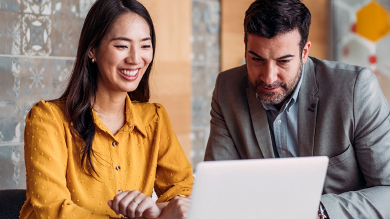 Diverse business professionals, an Asian woman and a man, smiling and collaborating intently on a laptop