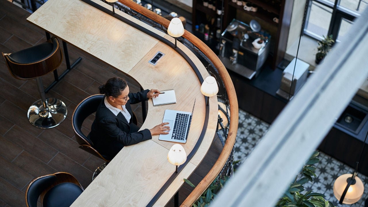 A woman sits at a desk with a laptop and a notebook with a pen poised over it. 