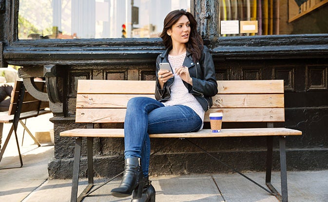 Woman sitting on a bench holding a phone and coffee cup nearby.